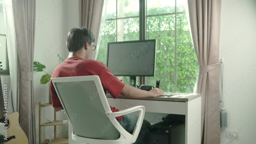 Asian man wear redshirt glasse sitting at home at the kitchen table, using tablet and watching changes in the currency exchange chart, looking at the computer monitor. freelancing.