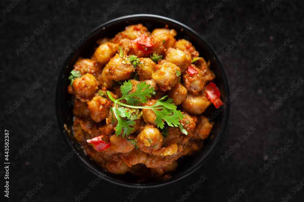 Chickpeas masala (Spicy chola or chhole curry) garnished with fresh green coriander and ingredients. Served in a black bowl. A Classic Indian typical Panjabi street food. Chickpeas masala (Spicy chola or chhole curry) garnished with fresh green coriander and ingredients. Served in a black bowl. A Classic Indian typical Panjabi street food.