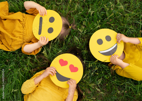 Фотография Three children lying on the grass are holding cardboard emoticons with different emotions in their hands: a sad, smiling happy smile, a loving smiley with hearts instead of eyes