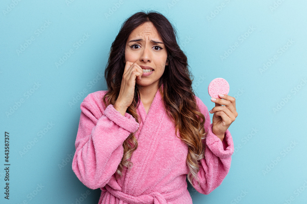 Young mexican woman wearing a bathrobe holding a make-up remover sponge isolated on blue background biting fingernails, nervous and very anxious. Young mexican woman wearing a bathrobe holding a make-up remover sponge isolated on blue background biting fingernails, nervous and very anxious.
