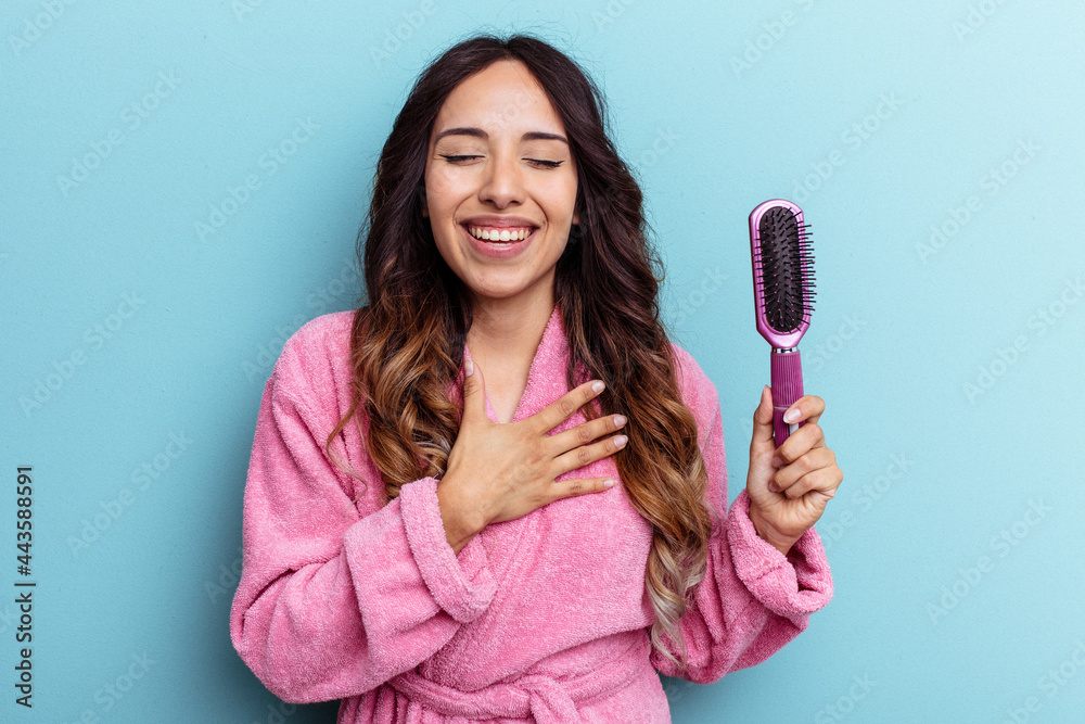 Young mexican woman wearing a bathrobe holding a brush isolated on blue background laughs out loudly keeping hand on chest. Young mexican woman wearing a bathrobe holding a brush isolated on blue background laughs out loudly keeping hand on chest.