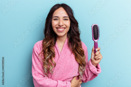 Young mexican woman wearing a bathrobe holding a brush isolated on blue background laughing and having fun.