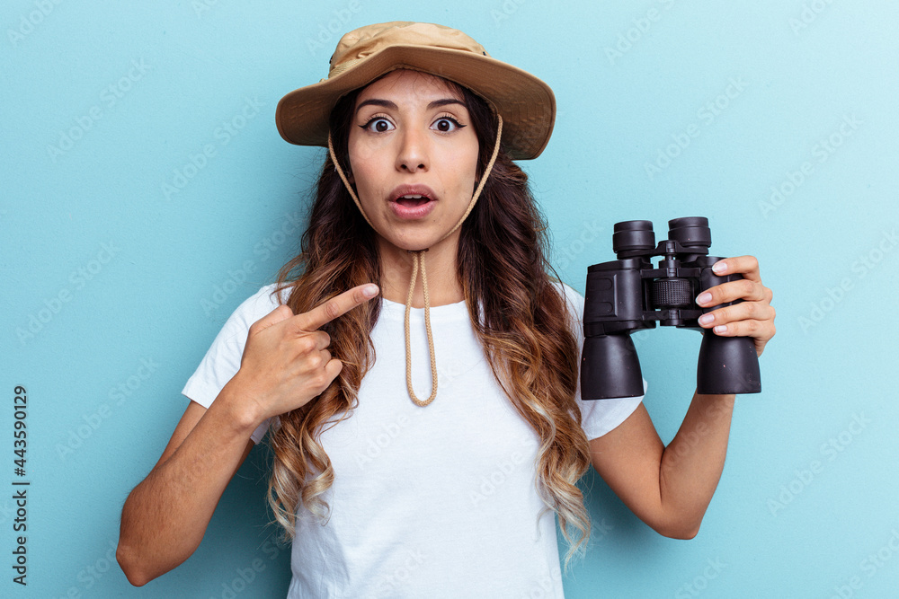 Young mexican woman holding binoculars isolated on blue background pointing to the side Young mexican woman holding binoculars isolated on blue background pointing to the side