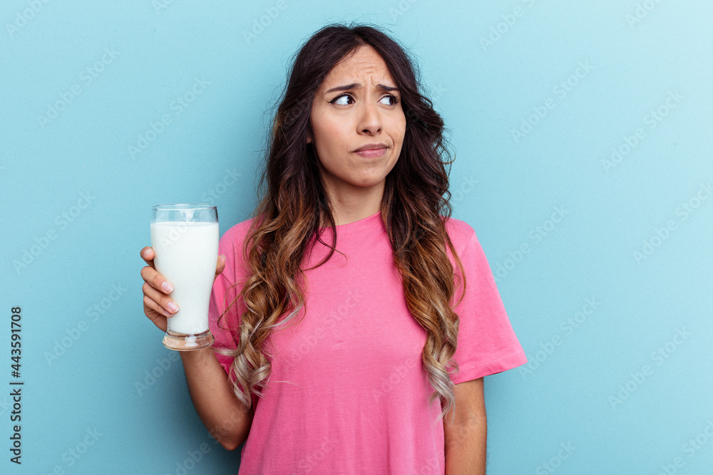 Young mixed race woman holding a glass of milk isolated on blue background confused, feels doubtful and unsure. Young mixed race woman holding a glass of milk isolated on blue background confused, feels doubtful and unsure.