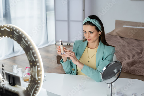 stylish woman pointing at face foundation near blurred phone holder with ring lamp.