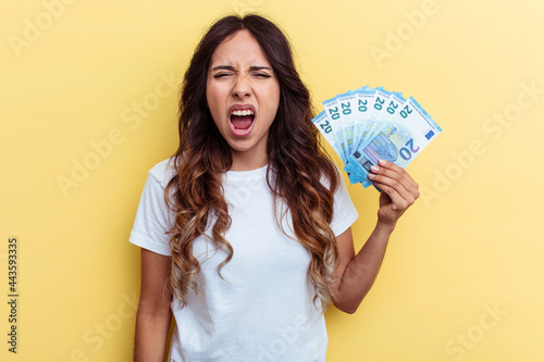 Young mixed race woman holding bills isolated on yellow background screaming very angry and aggressive.
