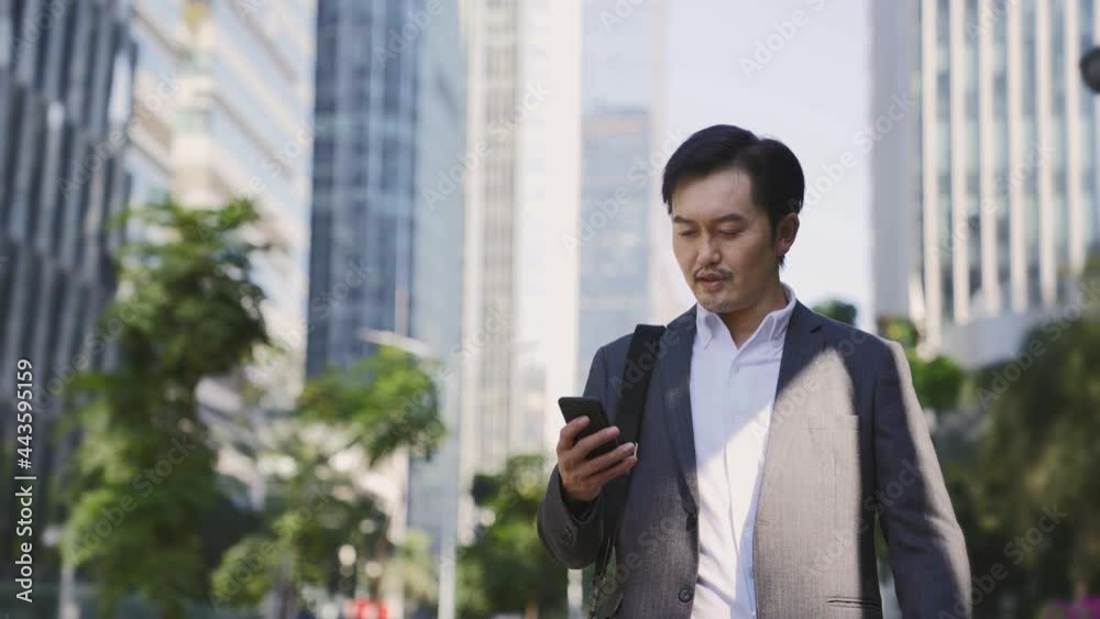 asian business man walking on street looking at cellphone in downtown financial district of modern city asian business man walking on street looking at cellphone in downtown financial district of modern city