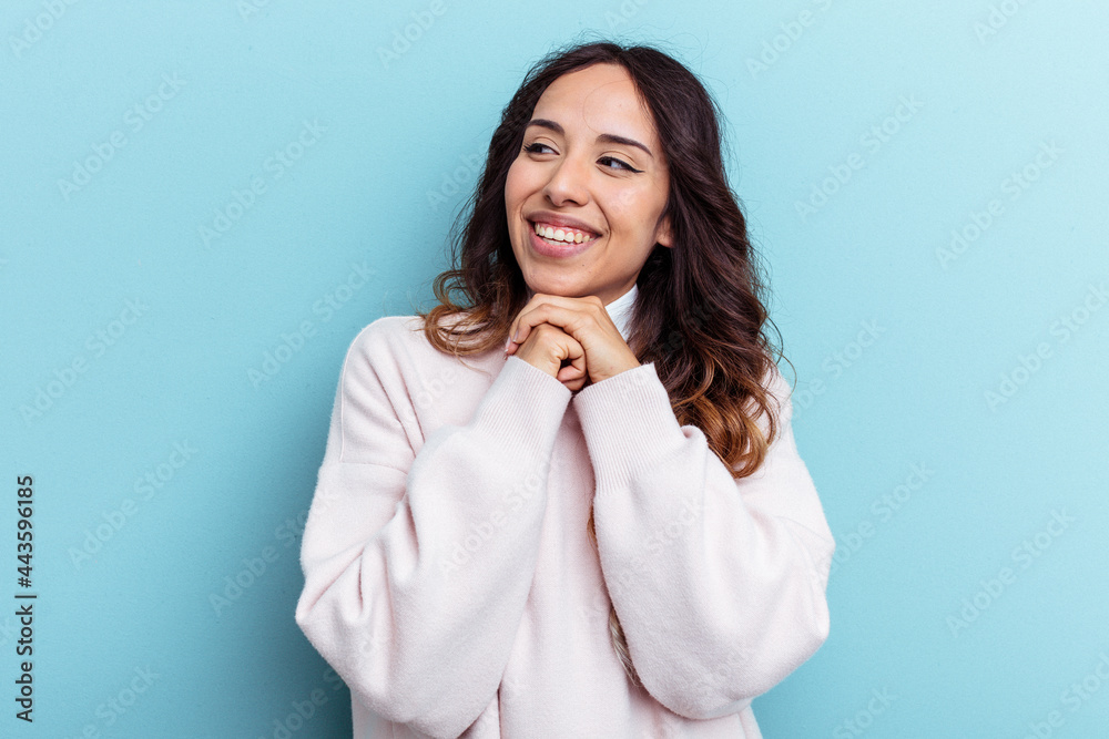 Young mexican woman isolated on blue background keeps hands under chin, is looking happily aside. Young mexican woman isolated on blue background keeps hands under chin, is looking happily aside.