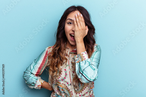 Young mexican woman isolated on blue background having fun covering half of face with palm.