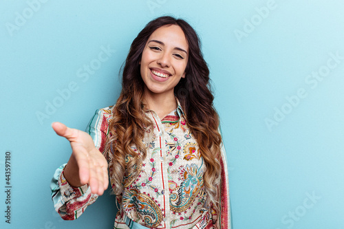 Young mexican woman isolated on blue background stretching hand at camera in greeting gesture.
