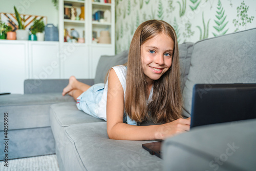 A little schoolgirl girl is studying at home, lying on the couch with a laptop. Distance learning online education