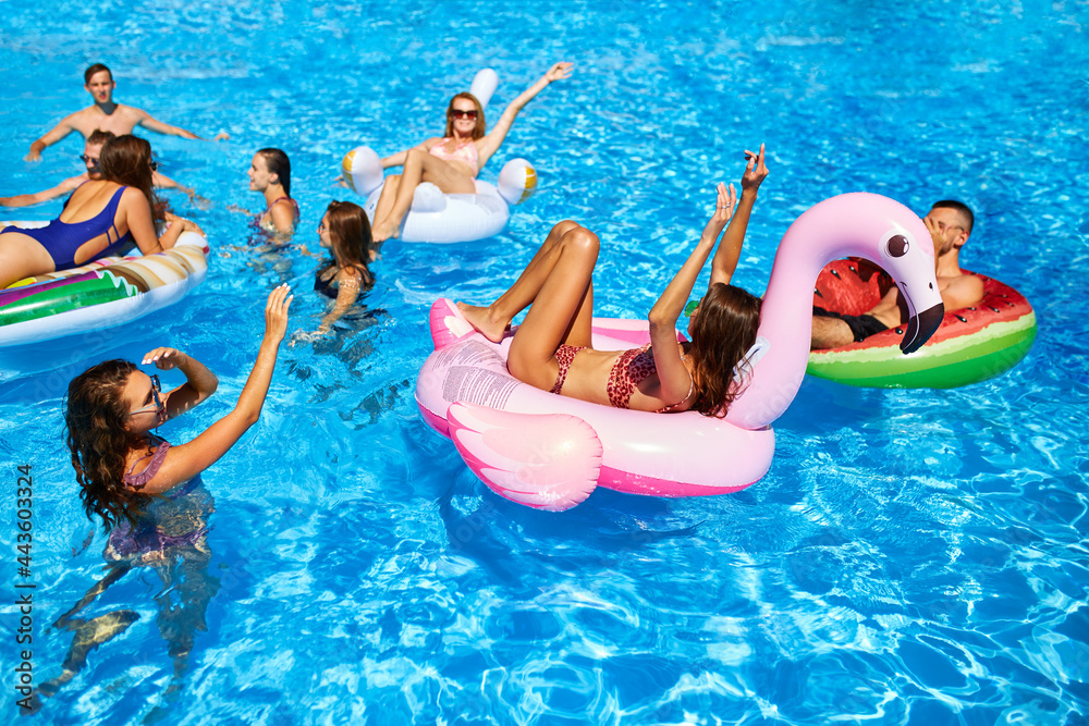 Friends having party in private holiday villa swimming pool. Happy young people swim on inflatable flamingo, swan at mattresses at luxury resort on sunny day. Girls in bikini lie in the sun.