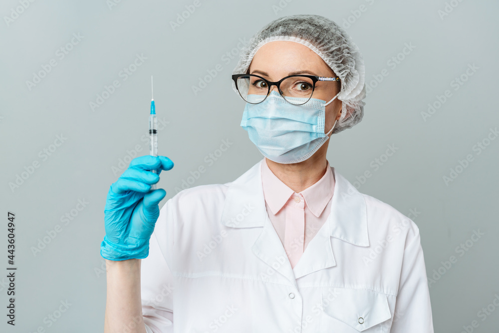 Female doctor or scientist in white medical gown, blue gloves, green cap and mask holds a syringe in hands on white background. She is ready to give an injection Female doctor or scientist in white medical gown, blue gloves, green cap and mask holds a syringe in hands on white background. She is ready to give an injection
