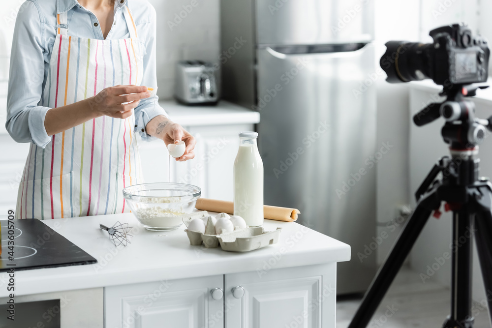 cropped view of woman breaking egg into bowl with flour near blurred digital camera.