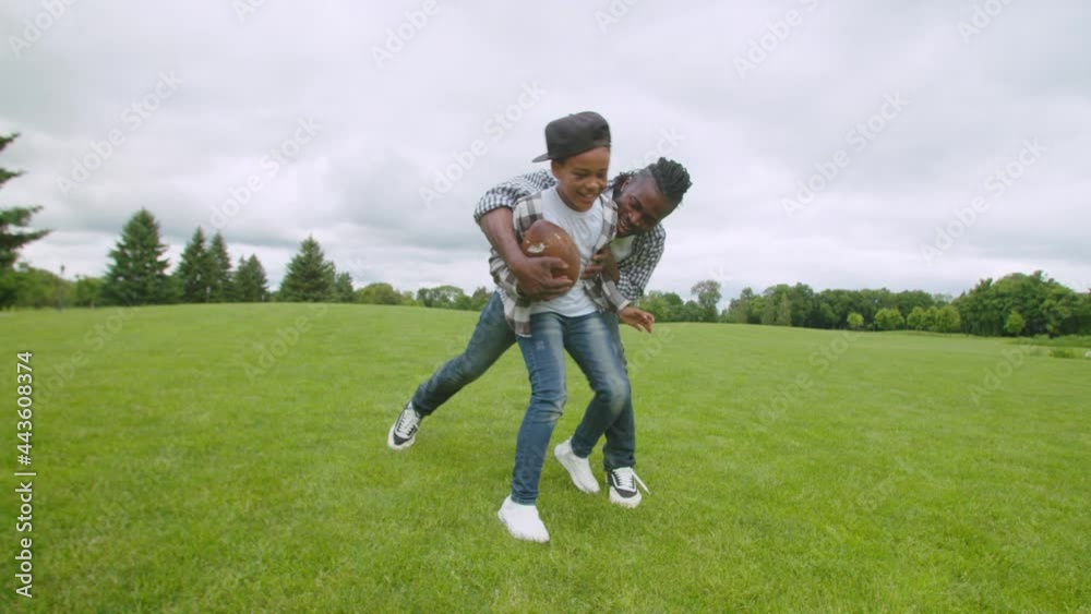 Carefree attractive black father and excited lovely school age boy fighting for ball possession, having fun, enjoying sport activities while family spending leisure playing american football game.