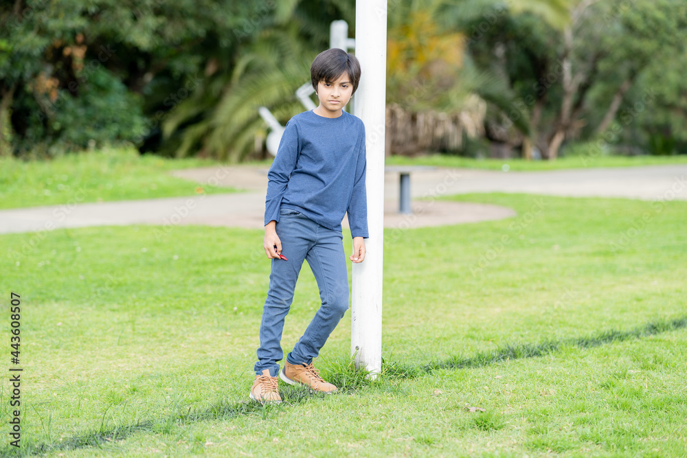Portrait of lonely child. Young boy standing in park leaning against pole looking towards camera. Shot in natural light. Portrait of lonely child. Young boy standing in park leaning against pole looking towards camera. Shot in natural light.