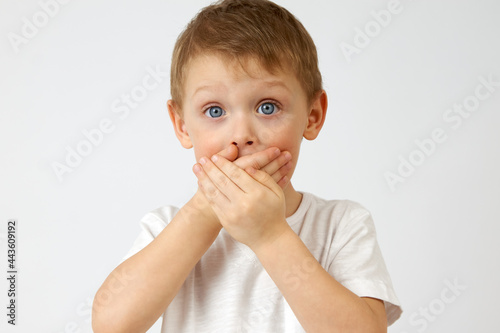 Child restrains emotion by covering his mouth with his hands, so as not to cry out from the horror that has seized him. Emotion of fear of a boy with wide open blue eyes on a white background