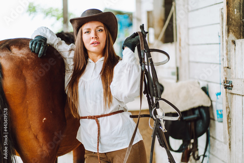 Young woman rider wearing a white shirt and hat with her brown horse in a stall, portrait.