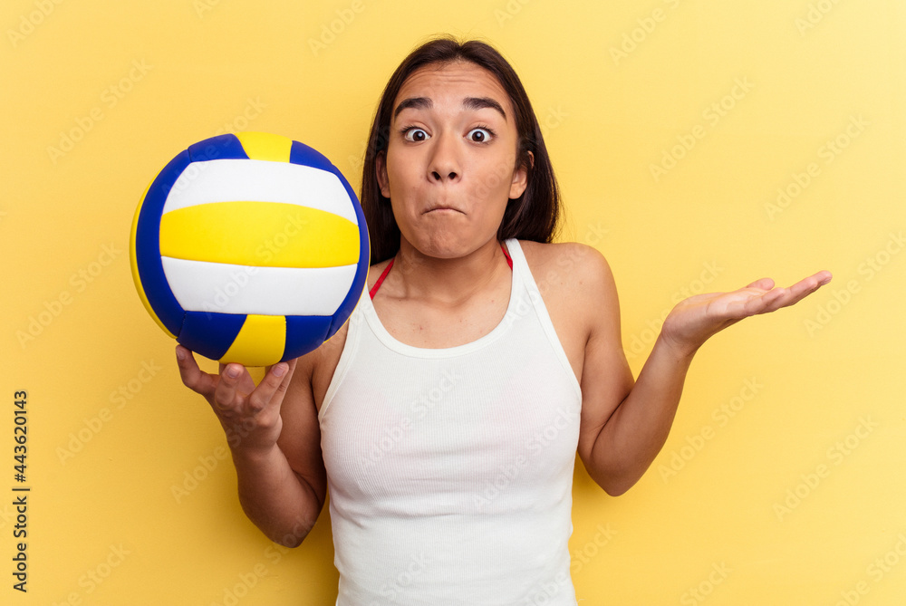 Young mixed race woman playing volleyball on the beach isolated on yellow background shrugs shoulders and open eyes confused.