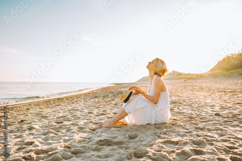 Young slim woman wearing white long dress and hat sit alone at the beach or ocean or sea. Copyspace.