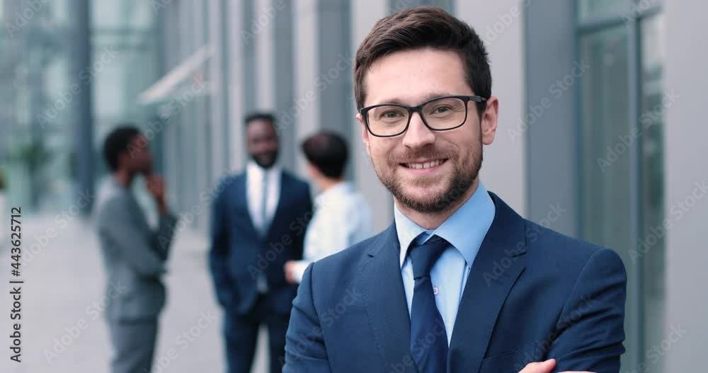 Confident and successful. Waist-up portrait of young elegant caucasian businessman is standing with crossed arms while looking at the camera with slight smile with his colleagues at the background Confident and successful. Waist-up portrait of young elegant caucasian businessman is standing with crossed arms while looking at the camera with slight smile with his colleagues at the background