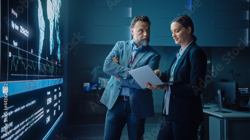 Project Manager and Computer Science Engineer Talk while Using Big Screen Display and a Laptop, Showing Infrastructure Infographics Data. Telecommunications Company System Control and Monitoring Room.