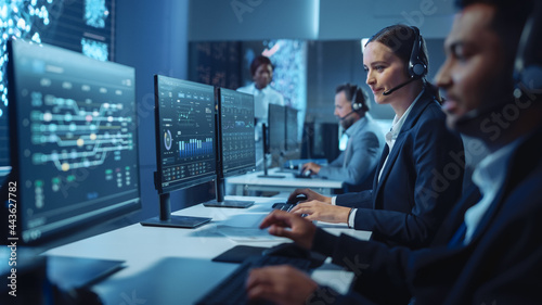 Foto Happy Confident Technical Customer Support Specialist Having a Headset Call while Working on a Computer in a Dark Monitoring and Control Room Filled with Colleagues and Display Screens