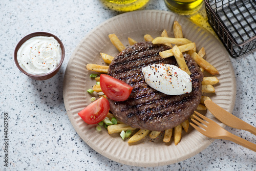 Serbian grilled pljeskavica with kaymak cheese, fries and red tomatoes on a carton plate, studio shot on a beige granite background