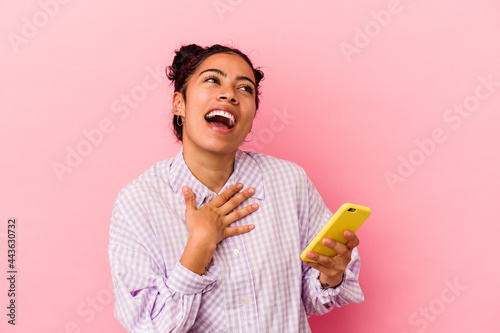 Young latin woman holding a mobile phone isolated on pink background laughs out loudly keeping hand on chest.