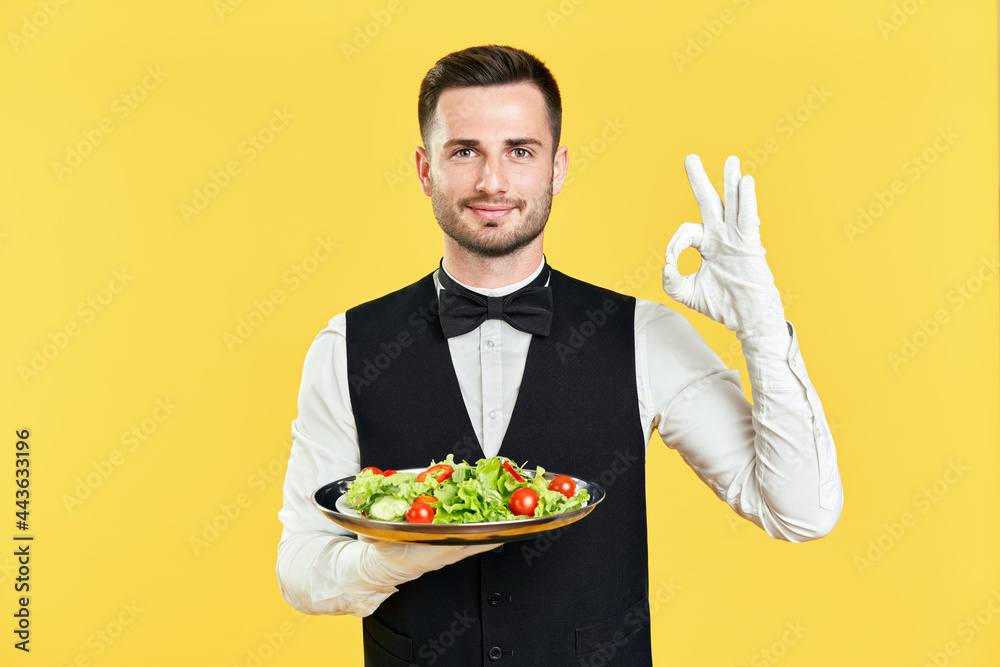 Happy smiling waiter holding plate with healthy vegetable salad doing ok sign with hand and fingers on yellow background Happy smiling waiter holding plate with healthy vegetable salad doing ok sign with hand and fingers on yellow background