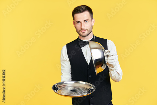 Elegant young waiter holding cloche over empty tray ready to serve on yellow background photo