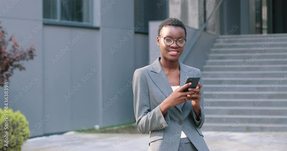 Attractive, adult multiracial woman in chic formal wear standing near the business center. She looking at camera and holding telephone with mail email in hands