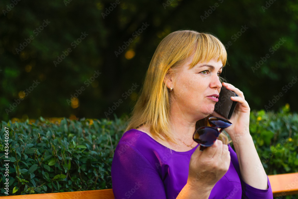 Defocus close-up caucasian blond woman talking, speaking on the phone outside, outdoor. 40s years old woman in purple blouse in park. Adult people using mobile phone. Copy space. Out of focus