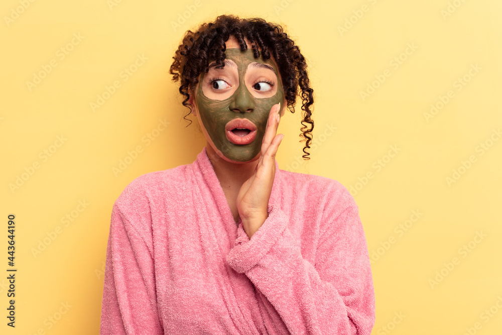 Young mixed race wearing facial mask isolated on yellow background is saying a secret hot braking news and looking aside