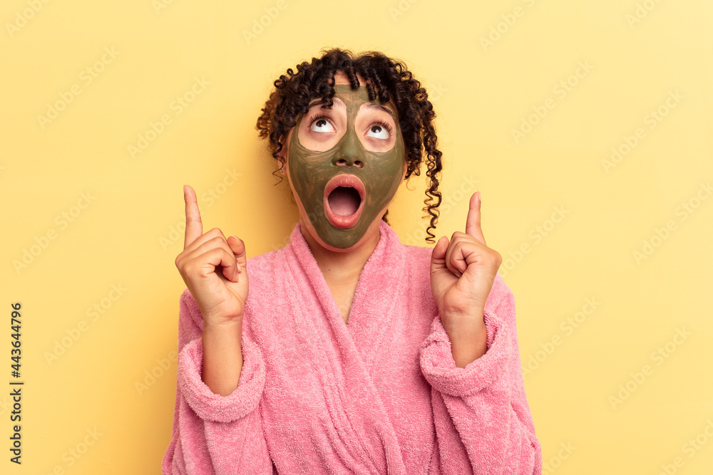 Young mixed race wearing facial mask isolated on yellow background pointing upside with opened mouth.