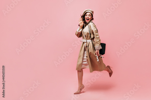 Girl in trench coat and beret happily waving her hand and running on pink background