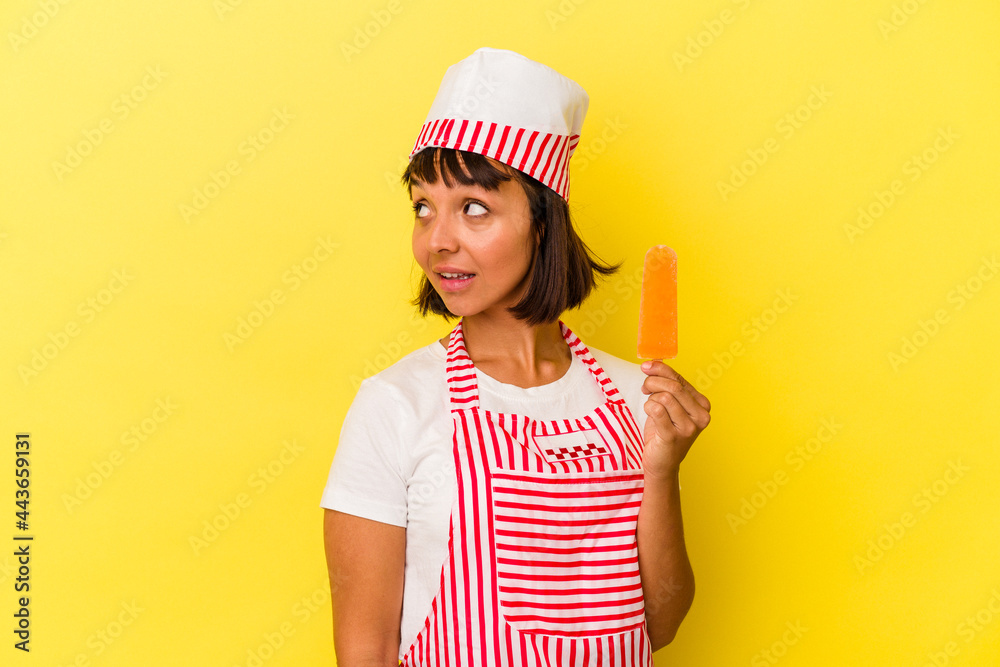 Young mixed race ice cream maker woman holding an ice cream isolated on yellow background looks aside smiling, cheerful and pleasant. Young mixed race ice cream maker woman holding an ice cream isolated on yellow background looks aside smiling, cheerful and pleasant.