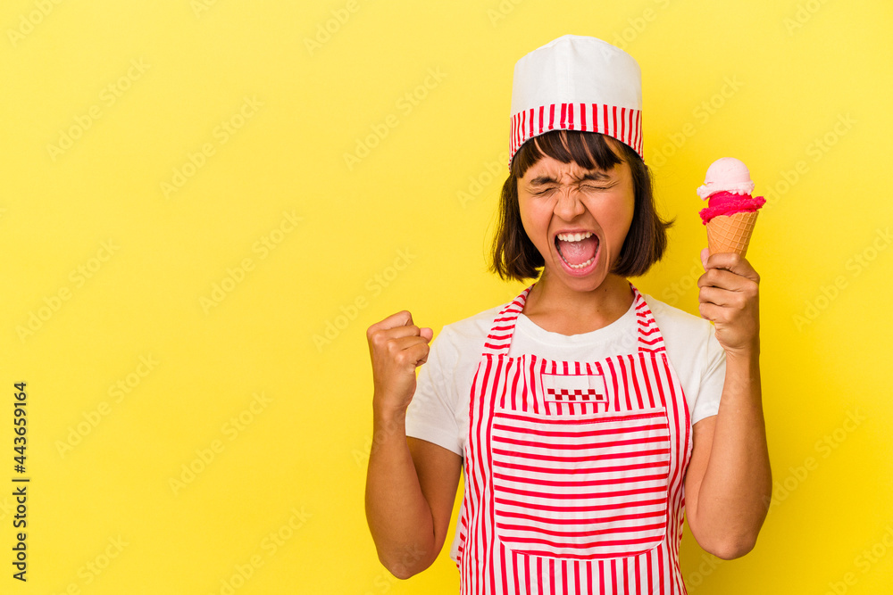 Young mixed race ice cream maker woman holding an ice cream isolated on yellow background raising fist after a victory, winner concept. Young mixed race ice cream maker woman holding an ice cream isolated on yellow background raising fist after a victory, winner concept.