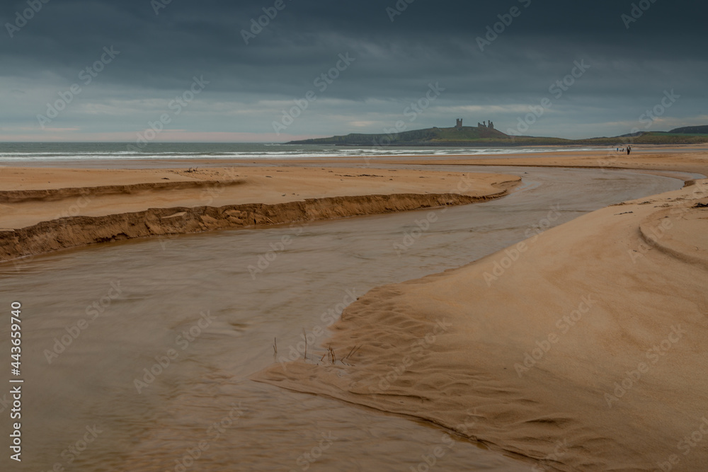 Dunstanburgh Castle in Northumberland, England, taken with the river meandering in the foreground & with cloudy skies overhead