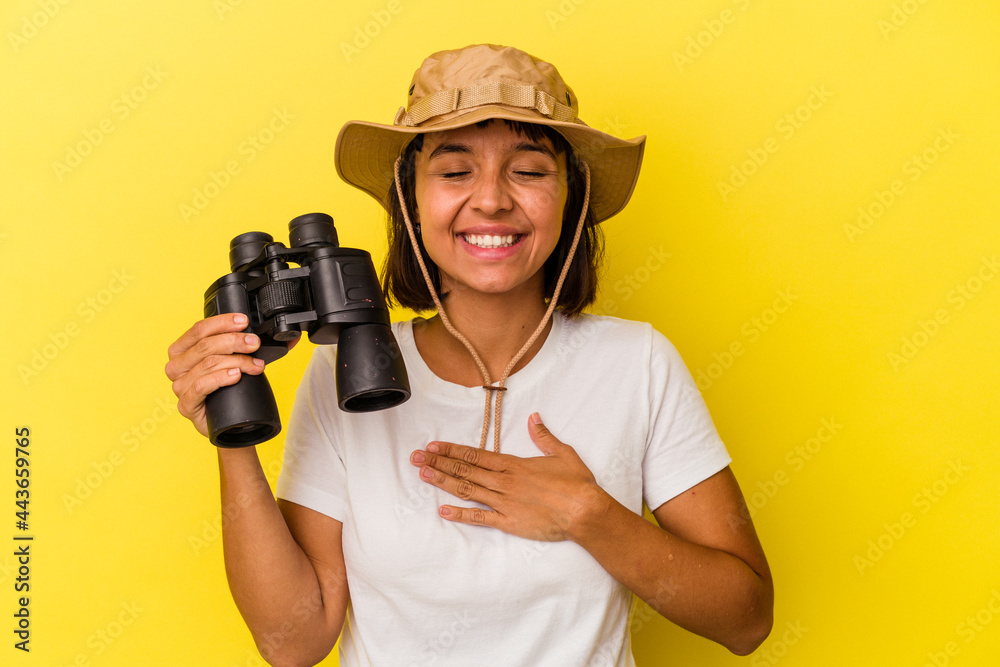 Young mixed race explorer woman holding binoculars isolated on yellow background laughs out loudly keeping hand on chest. Young mixed race explorer woman holding binoculars isolated on yellow background laughs out loudly keeping hand on chest.
