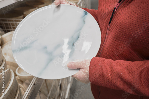 Caucasian woman in against viruses gloves buys large white plate with a marble pattern in a store. Concept of safe shopping and going to malls during epidemics and pandemics of viruses. Hands close up