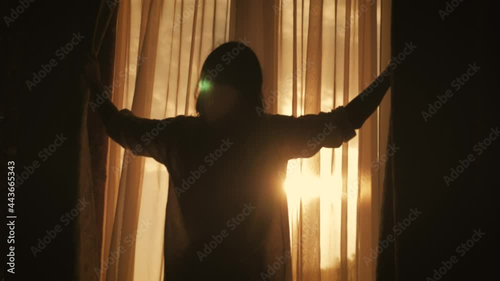 Lovely girl is awake and standing before window. Beautiful woman opening curtains and looking through the window.