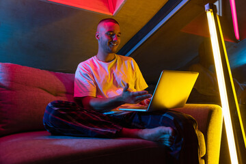 Portrait of handsome young Caucasian man sitting at home in neon orange pink lighted room. Stylish male model indoors. Reflection effect