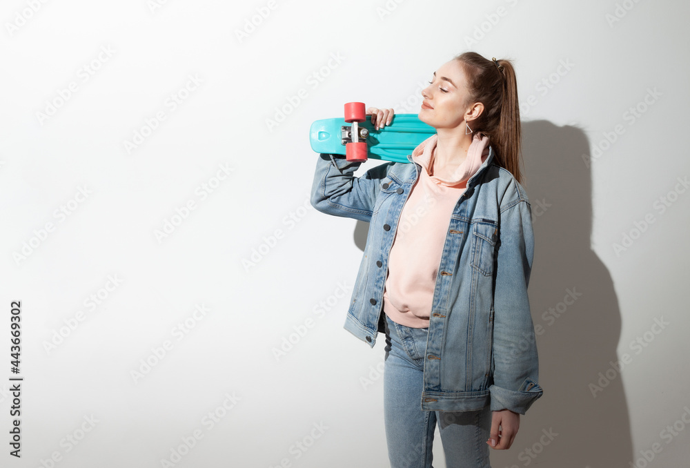 Young caucasian cheerful hipster woman in denim clothes with penny board. White background with long shadow Young caucasian cheerful hipster woman in denim clothes with penny board. White background with long shadow