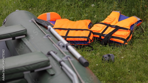 New orange life jackets on the green  grass on a summer day against the background of ainflatable boat with a paddle, safety when trip on water © Ilya