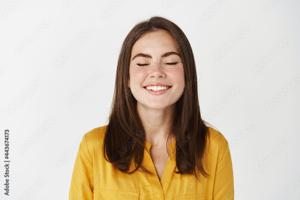 Close-up of happy and delighted young woman smiling with closed eyes, making a wish or dreaming, standing on white background Close-up of happy and delighted young woman smiling with closed eyes, making a wish or dreaming, standing on white background