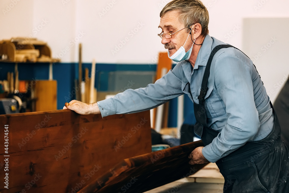 A gray-haired carpenter with glasses works with wood on a workbench in a carpentry shop. A pensioner in a medical mask at work. Employment and labor an elderly man in work clothes A gray-haired carpenter with glasses works with wood on a workbench in a carpentry shop. A pensioner in a medical mask at work. Employment and labor an elderly man in work clothes