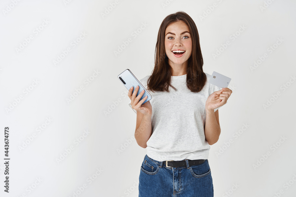 Ecommerce card payment. Happy young woman holding plastic credit card and smartphone, looking at camera surprised, standing over white background Ecommerce card payment. Happy young woman holding plastic credit card and smartphone, looking at camera surprised, standing over white background