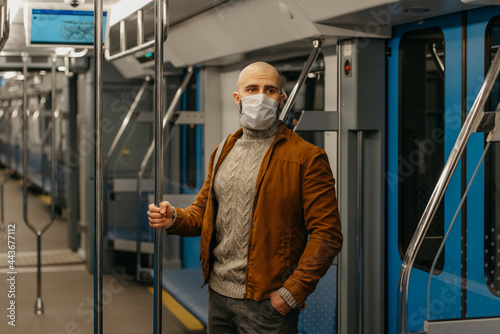 A man with a beard in a medical face mask to avoid the spread of COVID-19 is riding a subway car and holding the handrail. A bald guy in a surgical mask is keeping social distance on a train.