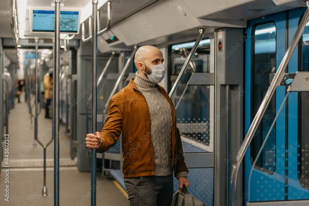 A man with a beard in a medical face mask to avoid the spread of coronavirus is riding a modern subway car. A bald guy in a surgical mask is keeping social distance on a train.
